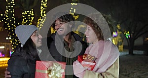 Excited happy friends holding gift boxes standing on garlands and lights background outdoor - New Year holidays