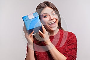 Excited happy girl in shaggy sweater holding gift box near ear, listening guess what`s inside, in anticipation