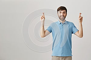 Excited happy good-looking male with beard standing with lifted hands and crossed fingers over gray background, desiring