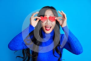 Excited happy woman holding two small paper hearts by her eyes isolated on blue background