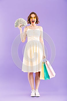 Excited happy woman isolated over purple wall background holding shopping bags and money.