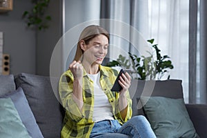 Excited and happy woman looking at phone screen, winning and good news, victory gesture, woman sitting on sofa at home