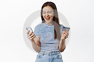 Excited happy woman scream from happiness, winning money cashback, holding credit card with mobile phone and rejoice