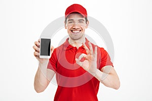 Excited happy young delivery man showing display of mobile phone make okay gesture.