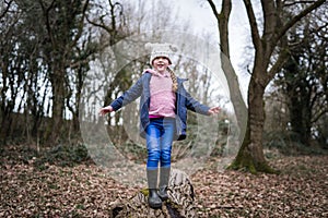 Excited happy young girl in wellington boots exploring countryside balancing on tree log with arms out exploring the outdoors