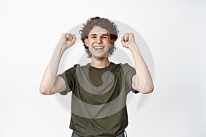 Excited happy young man cheering, raising hands up enthusiastic, winning and celebrating, standing in tshirt over white