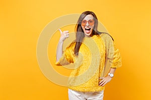 Excited happy young woman in fur sweater and heart orange glasses pointing thumb aside on copy space isolated on bright