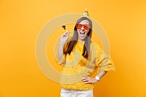 Excited happy young woman in orange funny eyeglasses birthday party hat with playing pipe celebrating isolated on bright