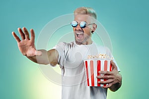 Excited hipster blonde man wearing stylish sunglasses and holding bucket with popcorn in studio in neon lights.