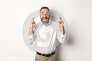 Excited and hopeful businessman making a wish, cross fingers and waiting, standing over white background