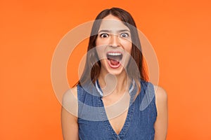 Excited impressed brunette woman in denim outfit standing with wide open mouth in surprise