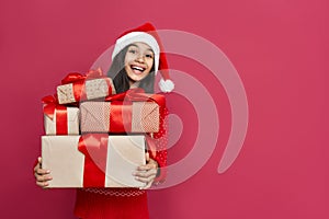 Excited indian kid girl wear santa hat hold many gifts boxes on red background.