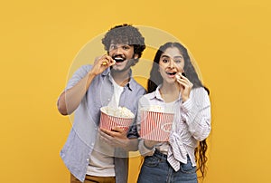 Excited indian man and woman eating popcorn at cinema, standing over yellow studio background