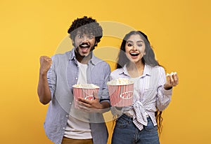 Excited indian man and woman eating popcorn, watching movie or football game, celebrating success of their team