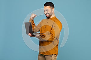 Excited italian man holding laptop pc computer doing winner gesture clench fist isolated on blue background