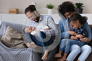 Excited joyful African parents cuddling, tickling kids on couch