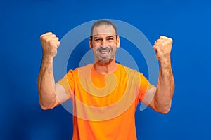 Excited joyful man in orange t-shirt standing isolated over blue background, celebrating success.