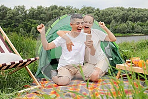 Excited joyful woman and man wearing casual clothing sitting at tent near the river, using mobile phone, clenched fists, rejoicing