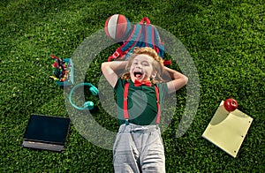 Excited kid learn online in virtual school using tablet on green grass outdoors, e-learning. Happy boy relaxing on the