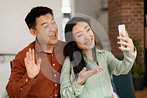 Excited korean couple using mobile phone, video calling in living room interior, smiling at cellphone webcamera