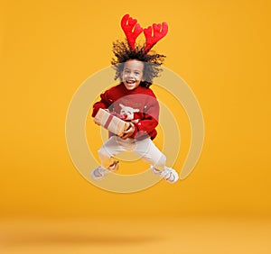 Excited little african american boy in Christmas sweater and deer antlers jumping up in air with Xmas gift box