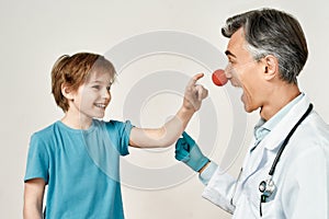 Excited little boy touching clown nose of friendly male pediatrician, isolated over grey background. Doctor and little