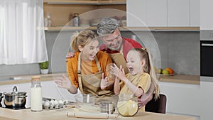 Excited little girl making flour splash and laughing, cooking dough together with loving parents at kitchen, empty space