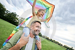 Excited little girl sitting on father`s shoulders and holding colorful kite in the beautiful green park on a warm summer