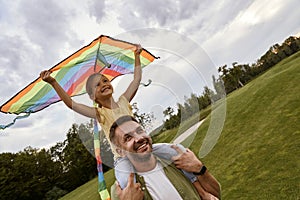 Excited little girl sitting on shoulders of her father and playing with colorful kite, daughter and daddy spending time