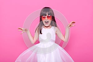 Excited little girl in white dress yelling with anger and frighten somebody, raising her hands, looking at camera, having dark