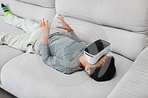 Excited little kid using virtual reality glasses. He lying on gray sofa and using new gadget technology