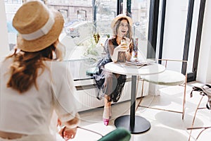 Excited long-haired girl in purple shoes telling friend last gossips while resting in cozy cafe. Indoor portrait of