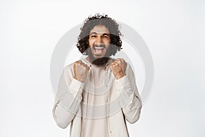 Excited lucky hispanic man winning, cheering with fist pumps and scream delighted, achieve goal, white background