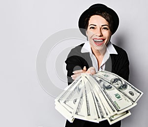 Excited lucky woman in stylish hat and black jacket is holding giving handing us a fan of dollars cash banknotes