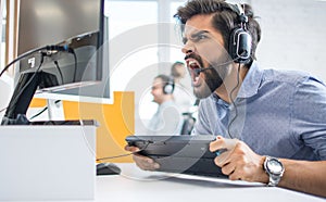Excited male customer support operator with headset shouting on computer monitor and holding keyboard at office