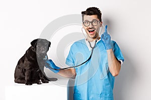 Excited male doctor veterinarian having an idea while examining cute pug dog with stethoscope, raising finger in eureka