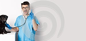 Excited male doctor veterinarian having an idea while examining cute pug dog with stethoscope, raising finger in eureka