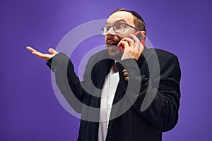 Excited man in glasses and yarmulke emotionally talking on phone, greeting with holiday against purple studio background