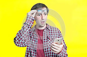 Excited man with open mouth in red checkered shirt taking off glasses on forehead and looking at smartphone