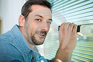 excited man spying through window blinds with binoculars