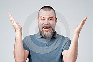 Excited mature man with beard in blue shirt standing with raised hands and looking at camera