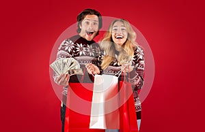 Excited millennial girl and her boyfriend holding shopping bags with Christmas gifts and money on red background