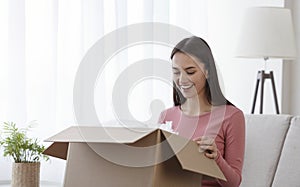 Excited millennial woman sitting on couch at home opening cardboard box