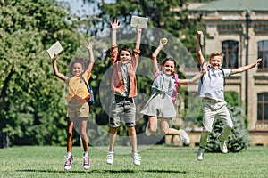 Excited multiethnic schoolkids jumping while holding books on lawn in park