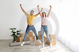 Excited multiracial couple celebrating relocation to their house, jumping among carton boxes on moving day, copy space