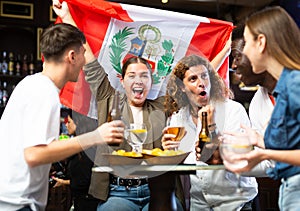 Excited multiracial fans with flag of Peru with pint of beer and snacks in the pub