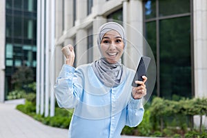 Excited muslim female cheering with mobile phone in raised hands while walking down business district. Emotional woman