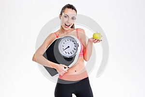 Excited positive fitness girl holding weighing scale and apple