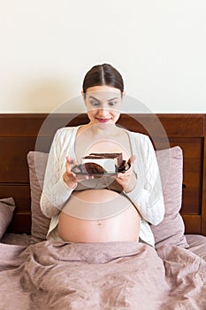 Excited pregnant woman is eating a slice of cake resting in bed at home. Love to sweet during pregnancy