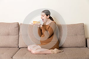 Excited pregnant woman is eating a slice of cake resting on the sofa at home. Love to sweet during pregnancy